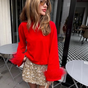 Pode incluir: Uma blusa vermelha de manga comprida com punhos largos adornados com penas vermelhas a condizer, combinada com uma saia curta coberta de lantejoulas douradas circulares brilhantes. Uma mala branca com pega estruturada é segurada em baixo.