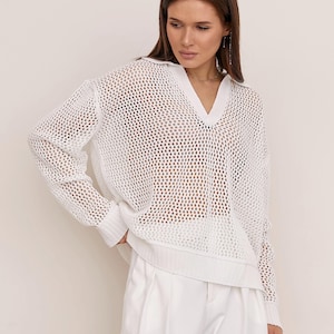 May include: A white long-sleeve sweater featuring an open-knit mesh construction, a collared V-neckline, and ribbed cuffs and hem. The garment is paired with white trousers, creating a monochromatic look with a textured, semi-sheer finish.