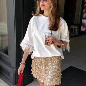 Puede incluir: Una camiseta blanca con adornos de plumas en las mangas y una minifalda dorada con lentejuelas. La persona lleva gafas de sol y sostiene un bolso rojo y una bebida en un vaso transparente.