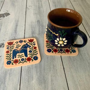 K&ouml;nnte beinhalten: Zwei quadratische Untersetzer mit einem Volkskunstdesign mit einem blauen Pferd und floralen Elementen in Rot, Blau und Gr&uuml;n auf pfirsichfarbenem Hintergrund. Eine dunkelblaue Tasse mit wei&szlig;en floralen Designs steht auf einem Untersetzer.