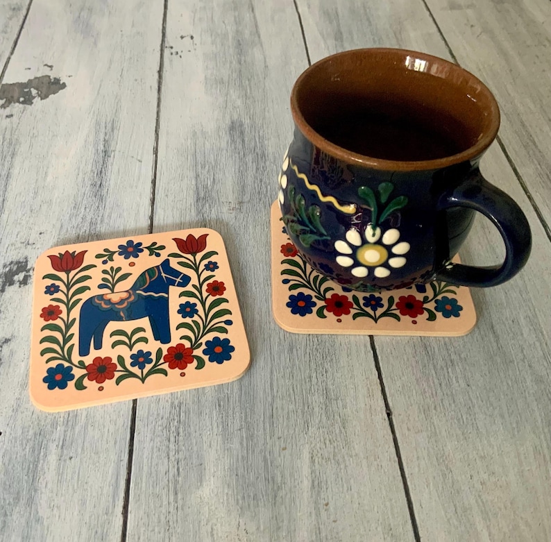 K&ouml;nnte beinhalten: Zwei quadratische Untersetzer mit einem Volkskunstdesign mit einem blauen Pferd und floralen Elementen in Rot, Blau und Gr&uuml;n auf pfirsichfarbenem Hintergrund. Eine dunkelblaue Tasse mit wei&szlig;en floralen Designs steht auf einem Untersetzer.