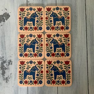 K&ouml;nnte beinhalten: Set mit sechs quadratischen Untersetzern mit einem Volkskunst-Design. Jeder Untersetzer zeigt ein blaues Pferd in einem floralen Rahmen mit roten, blauen und gr&uuml;nen Akzenten auf hellpfirsichfarbenem Hintergrund. Hergestellt aus strapazierf&auml;higem Material.