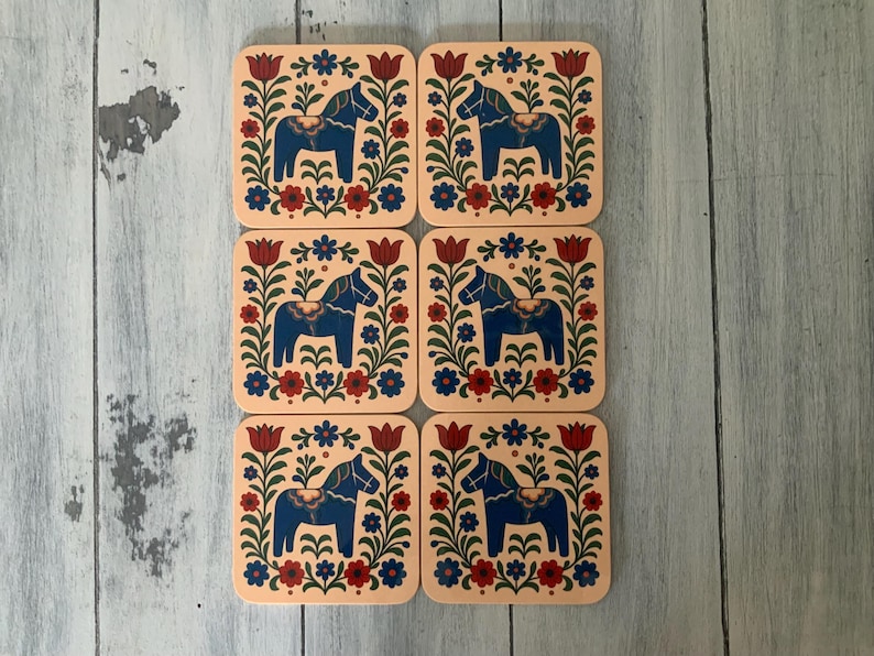 K&ouml;nnte beinhalten: Set mit sechs quadratischen Untersetzern mit einem Volkskunst-Design. Jeder Untersetzer zeigt ein blaues Pferd in einem floralen Rahmen mit roten, blauen und gr&uuml;nen Akzenten auf hellpfirsichfarbenem Hintergrund. Hergestellt aus strapazierf&auml;higem Material.