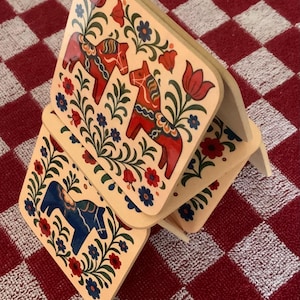 K&ouml;nnte beinhalten: Holzuntersetzer mit einem Volkskunst-Design. Jeder Untersetzer zeigt ein Dala-Pferd in Rot oder Blau, umgeben von floralen Mustern in Rot, Blau und Gr&uuml;n. Die Untersetzer sind auf einer rot-wei&szlig; karierten Oberfl&auml;che gestapelt.