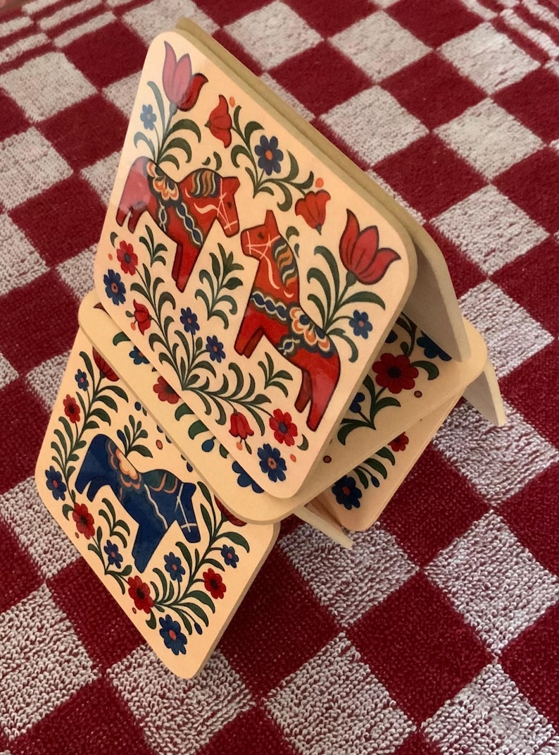 K&ouml;nnte beinhalten: Holzuntersetzer mit einem Volkskunst-Design. Jeder Untersetzer zeigt ein Dala-Pferd in Rot oder Blau, umgeben von floralen Mustern in Rot, Blau und Gr&uuml;n. Die Untersetzer sind auf einer rot-wei&szlig; karierten Oberfl&auml;che gestapelt.