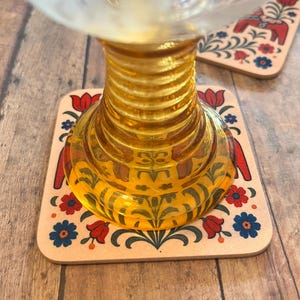 K&ouml;nnte beinhalten: Ein klares Glas steht auf einem dekorativen Untersetzer. Der Untersetzer hat ein Blumendesign mit roten Tulpen, blauen Blumen und einem roten Pferd. Das Glas hat einen gerippten, bernsteinfarbenen Stiel. Der Untersetzer ist quadratisch.