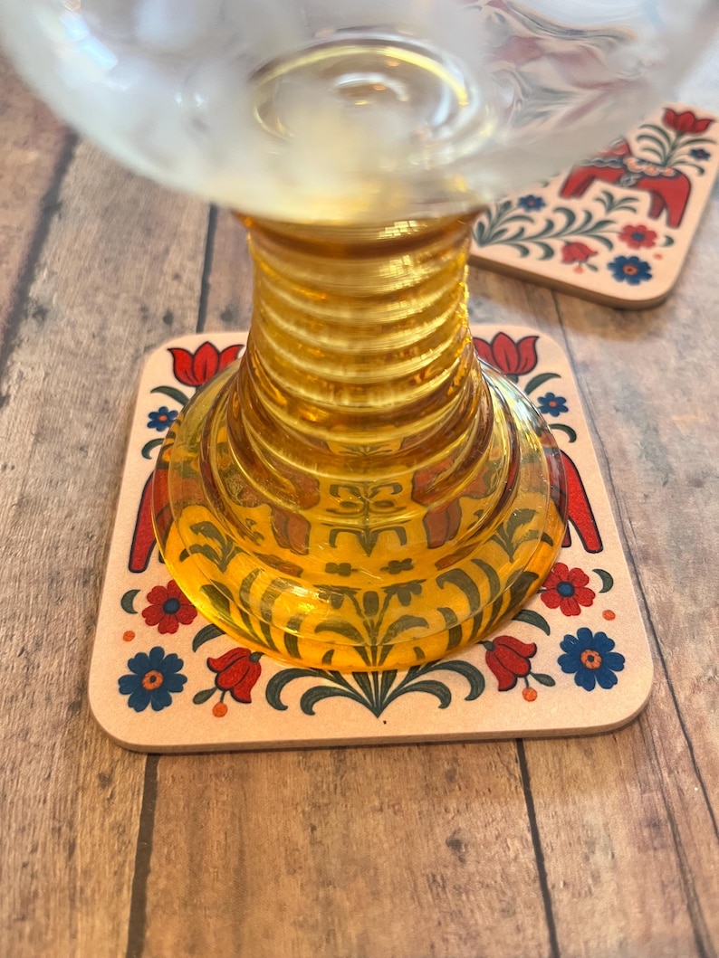 K&ouml;nnte beinhalten: Ein klares Glas steht auf einem dekorativen Untersetzer. Der Untersetzer hat ein Blumendesign mit roten Tulpen, blauen Blumen und einem roten Pferd. Das Glas hat einen gerippten, bernsteinfarbenen Stiel. Der Untersetzer ist quadratisch.