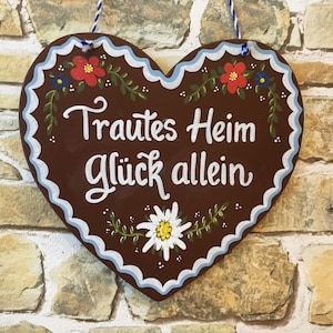 May include: A brown heart-shaped wooden sign with white lettering that reads "Trautes Heim Glück allein". The sign is decorated with blue and white trim, red and white flowers, and a white edelweiss flower.