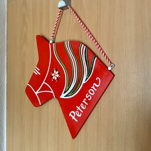 Może przedstawiać: Drewniany czerwony koń Dala z białymi i zielonymi detalami malowanymi, wiszący na drzwiach z białym hakiem. Koń ma napisane na sobie imię "Peterson".