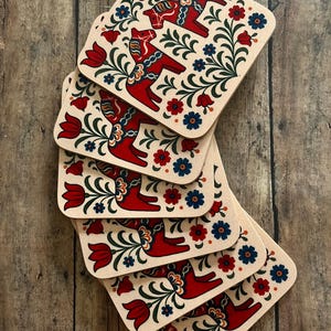 K&ouml;nnte beinhalten: Ein Stapel quadratischer Untersetzer mit beigem Hintergrund. Jeder Untersetzer zeigt ein rotes Dala-Pferd-Design, umgeben von roten Tulpen, blauen und roten Blumen und gr&uuml;nem Laub. Die Untersetzer sind auf einer Holzoberfl&auml;che angeordnet.