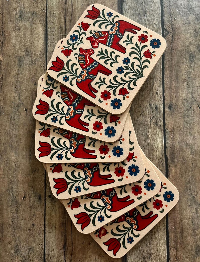 K&ouml;nnte beinhalten: Ein Stapel quadratischer Untersetzer mit beigem Hintergrund. Jeder Untersetzer zeigt ein rotes Dala-Pferd-Design, umgeben von roten Tulpen, blauen und roten Blumen und gr&uuml;nem Laub. Die Untersetzer sind auf einer Holzoberfl&auml;che angeordnet.