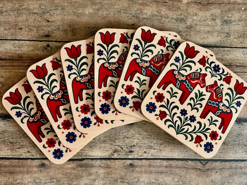 K&ouml;nnte beinhalten: Set aus f&uuml;nf quadratischen Holzuntersetzern mit traditionellem Dala-Pferdemotiv. Jeder Untersetzer zeigt ein rotes Pferd, rote Tulpen und blaue und rote florale Akzente auf beigem Hintergrund.