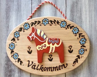 May include: A wooden oval sign with a painted Dala horse and the word "Välkommen" in Swedish, which means "Welcome". The sign is decorated with blue flowers and green leaves.