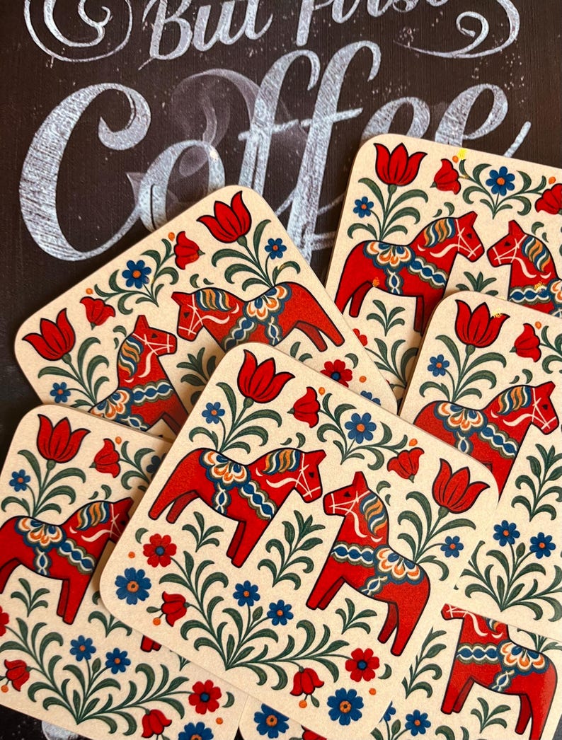 K&ouml;nnte beinhalten: Eine Sammlung cremefarbener Untersetzer mit einem traditionellen schwedischen Dala-Pferdemotiv. Die roten Pferde sind von blauen und gr&uuml;nen Blumenmustern umgeben. Im Hintergrund steht in Wei&szlig; "But First Coffee".