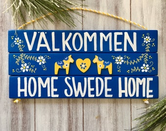 May include: A blue wooden sign with the text "Välkommen Home Swede Home" in white paint. The sign is decorated with yellow and white flowers, green leaves, and two yellow and blue Dala horses.