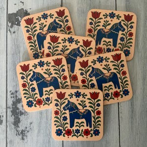 K&ouml;nnte beinhalten: Sechs quadratische Untersetzer mit hellpfirsichfarbenem Hintergrund. Jeder Untersetzer zeigt ein blaues Dala-Pferd-Design, umgeben von roten Tulpen, blauen und roten Blumen und gr&uuml;nen Bl&auml;ttern. Die Untersetzer liegen auf einer hellblauen Holzoberfl&auml;che.