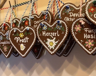 Op de afbeelding: Een verzameling hartvormige peperkoekkoekjes versierd met witte glazuur en kleurrijke bloemen. De koekjes hebben verschillende Duitse zinnen erop geschreven, waaronder "Grüß gott", "Engelche", "Prost", "Herzerl", "Devine Oktober", "Devine Oktoberf 2024" en "Fürs Hasi".