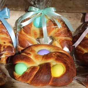 May include: Braided Easter bread, some with coloured eggs, wrapped in cellophane and tied with ribbons. Ribbons are in blue, pink, and green. The bread is a golden brown colour, with a braided design.