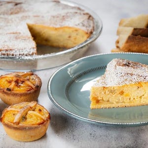 May include: A selection of baked goods, including a large pie with a slice removed, dusted with powdered sugar. A slice of pie sits on a blue plate, and several small tarts with fruit toppings are also visible. Sliced bread is in the background.