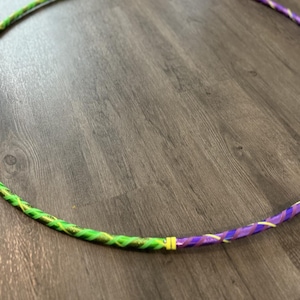 May include: A green and purple hula hoop with a yellow stripe. The hoop is made of plastic and has a shiny finish.