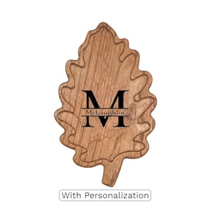 Puede incluir: Una tabla de cortar de madera con forma de hoja con un borde tallado y una inicial y un nombre personalizados. La tabla es marrón con una inicial oscura "M" y el nombre "McLaughlin" en negro. El texto "Con personalización" está en la parte inferior.
