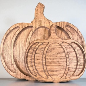 Puede incluir: Dos decoraciones de madera con forma de calabaza. Las calabazas están talladas en madera de color marrón claro, con ranuras detalladas para representar los segmentos de la calabaza. La calabaza más grande está detrás de la más pequeña, creando un efecto de capas. La veta de la madera es visible.