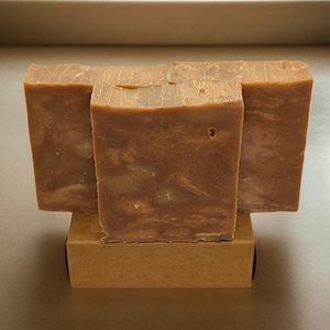 Puede incluir: Tres barras de jabón marrón con remolinos de blanco y beige, apiladas una encima de la otra. Las barras de jabón están sobre una caja de cartón marrón.