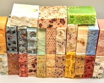 May include: A collection of rectangular soap bars arranged in a grid, featuring various colors including white, green, orange, pink, and blue. The bars display diverse textures, some with embedded botanical pieces, honeycomb patterns, and swirls.