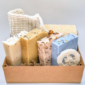 Puede incluir: Una cesta de regalo llena de productos de baño naturales, que incluyen cuatro barras de jabón, una esponja de lufa y un puff de baño de malla. Los jabones son en tonos beige, rosa y azul, y la cesta es de cartón marrón.
