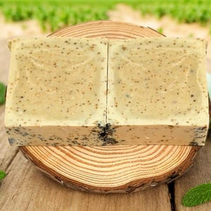 May include: Two bars of handmade soap on a wooden slice. The soap is light green and has a speckled appearance. The soap is surrounded by green leaves.