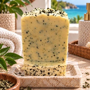 May include: A rectangular bar of pale yellow soap with visible dark green herbal flecks throughout the surface. The soap is positioned upright on a square, light-colored, textured stone soap dish, showcasing a smooth yet speckled texture.