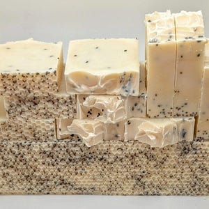 Puede incluir: Una pila de barras de jabón artesanales con un diseño de panal. Las barras de jabón son de color beige claro con motas negras. Las barras de jabón están apiladas una encima de la otra, siendo la barra inferior la más grande.