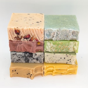 Puede incluir: Una pila de ocho barras de jabón artesanales en varios colores y patrones. Las barras están dispuestas en dos filas de cuatro, con la fila superior que presenta una barra beige claro con flores secas, una barra verde con un patrón moteado, una barra rosa con un patrón de remolino y una barra blanco y negro con un patrón de remolino. La fila inferior presenta una barra amarilla con semillas negras, una barra amarilla con un patrón moteado, una barra beige con semillas negras y una barra amarilla con un patrón moteado.