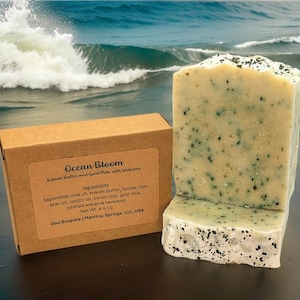Op de afbeelding: Een Ocean Bloom zeep met een lichtgroene en witte kleur. De zeep is gemaakt met kokumboter, geitenmelk en wakame zeewier. De zeep is verpakt in een bruine doos met de ingrediënten op de zijkant. De zeep weegt 4-5 ounce.
