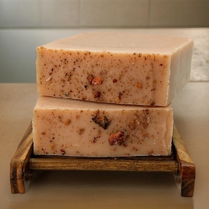 May include: Two bars of pink soap with brown flecks and pink flower petals on a wooden soap dish.