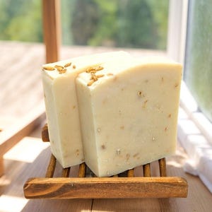 Puede incluir: Dos barras de jabón blanco con copos de avena en un jabonera de madera. El jabón está sobre una superficie de madera cerca de una ventana.