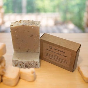 Puede incluir: Dos barras de jabón de avena con una caja de cartón marrón. La caja tiene el texto "Owl Soapery, Nature's Beauty, Artfully Crafted.  Avocado", y "Handmade Soaps, CO, USA".