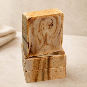 Peut inclure: Une pile de trois pains de savon rectangulaires avec un motif marbré dans des tons beige, crème et brun foncé. La surface du savon présente un aspect texturé et moucheté avec des taches visibles dans les tourbillons de couleurs claires et foncées.