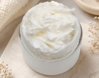 May include: A small, round, frosted glass jar filled with a thick, white, whipped cream-like substance. The surface of the white cream has visible peaks and swirls, indicating a dense, smooth texture contained within the clear, open-top vessel.