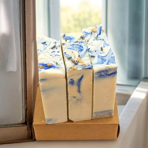 Puede incluir: Tres barras de jabón artesanal en una caja de cartón. El jabón es blanco con remolinos azules y motas de lavanda seca.