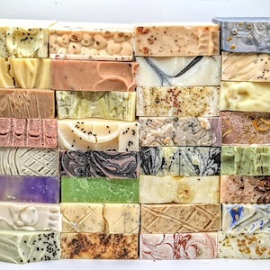 Puede incluir: Una pila de 24 barras de jabón hechas a mano en varios colores y patrones. Los jabones están dispuestos en un patrón de cuadrícula y presentan una variedad de diseños, que incluyen remolinos, rayas y motas.
