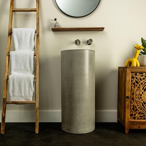 May include: A gray concrete pedestal sink with a round top and a chrome faucet. The sink is in a bathroom with a wooden ladder, a shelf, and a small wooden cabinet with a carved door.