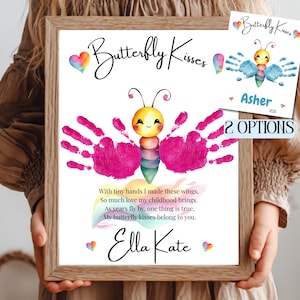 Op de afbeelding: Een ingelijste print met een vlinderontwerp gemaakt van handafdrukken. De vlinder heeft een gele kop en een kleurrijk lichaam. De tekst "Butterfly Kisses" staat bovenaan, met de naam "Ella Kate" onderaan. Een kleinere print toont een vergelijkbaar ontwerp met de naam "Asher".