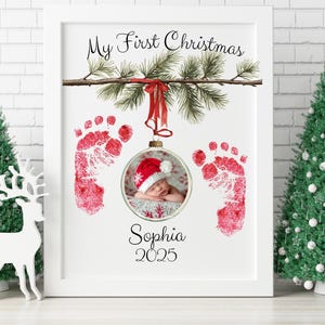 May include: A framed print with the text "My First Christmas" and a baby's photo inside a Christmas ornament. Red footprints flank the ornament. The baby is wearing a Santa hat. The name "Sophia" and the year "2025" are below the ornament.