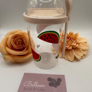 May include: A clear plastic cup with a watermelon design, a matching lid, and a spoon. The cup has a light pink rim and a clear dome lid. A business card with the text "Balloon Bliss by Lisa" is in the foreground. Peach-coloured flowers are in the background.