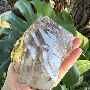 May include: A large, translucent smoky quartz crystal cluster with a jagged, multi-faceted surface. The mineral displays varying shades of brown and gray, featuring sharp, angular edges and natural internal fractures that catch the light.