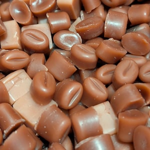 May include: Close-up of a pile of assorted brown candies. Some candies are shaped like coffee beans, while others are square or rectangular. The candies vary in shade from light tan to dark brown, with a glossy finish.