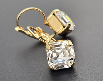 May include: A pair of gold-colored earrings with square, clear crystal pendants. The earrings have a lever-back closure and are set against a dark gray background. The crystals reflect light, creating a sparkling effect.