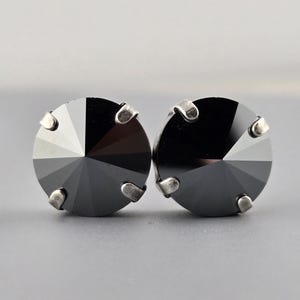 May include: A pair of round, faceted black crystal stud earrings. Each earring is divided into sections of varying shades of gray and black, with silver-toned prongs securing the crystal. The earrings are set against a gray background.