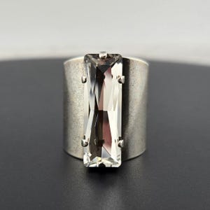 May include: A silver ring with a large, clear rectangular crystal set in the center. The ring is wide and has a textured finish.